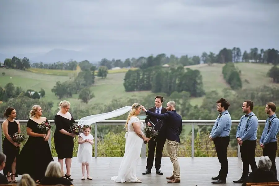 Wedding party stands on a deck overlooking rolling hills as the bride arrives.