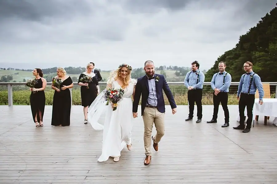 Newlyweds walk hand in hand across the deck in front of their wedding party.