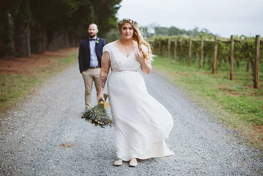 Bride walking along a gravel road with the groom behind her.
