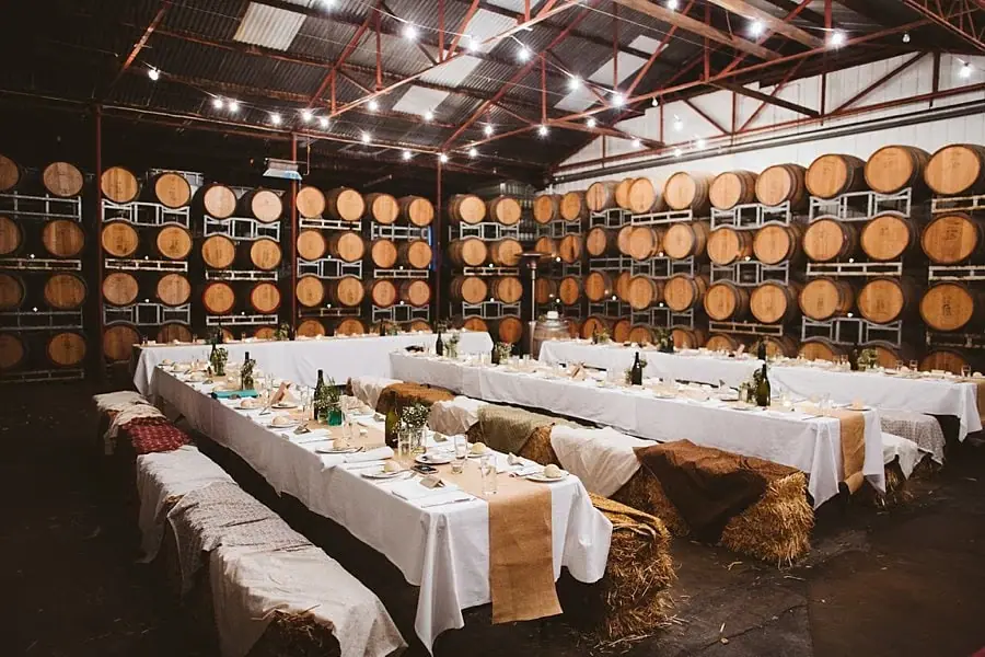 Reception tables with white cloths and hay-bale seating surrounded by stacked wine barrels.