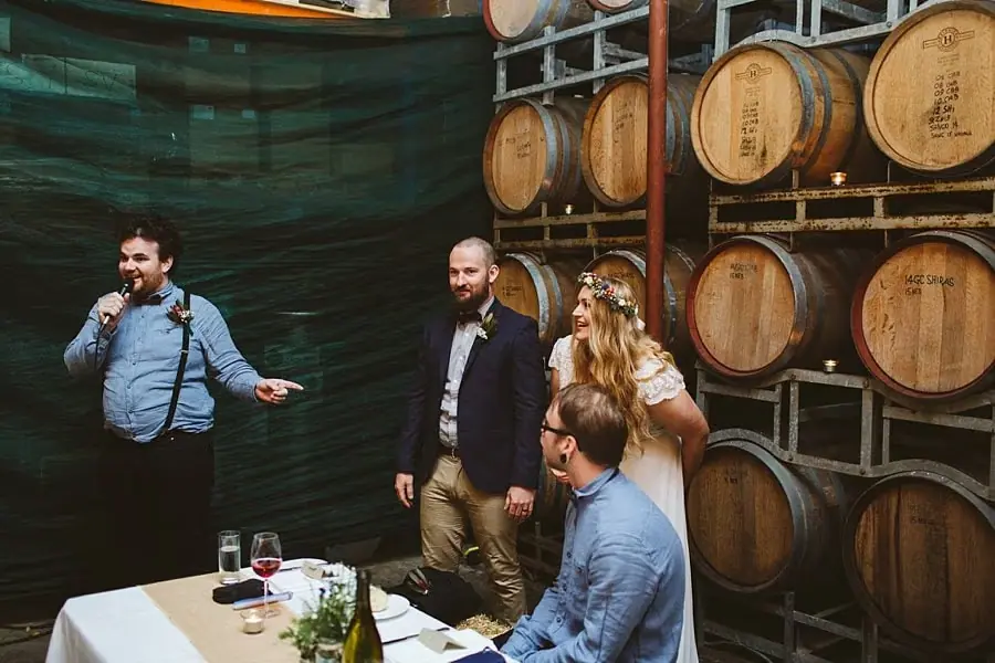 Man speaking into a microphone beside the bride and groom in front of wine barrels.