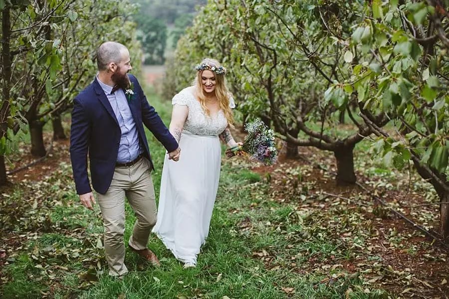 Bride and groom walking hand in hand between rows of trees.
