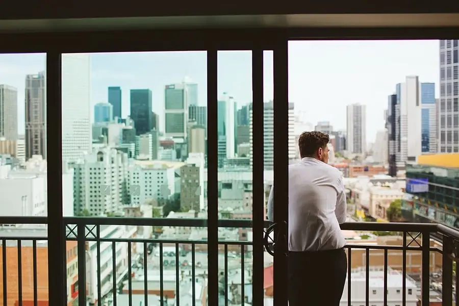 Man in a white shirt standing on a balcony overlooking city buildings.
