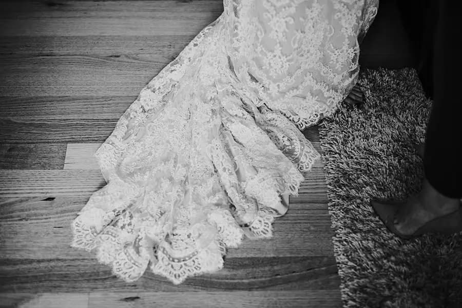 Black-and-white detail of a lace wedding dress train on a wooden floor.
