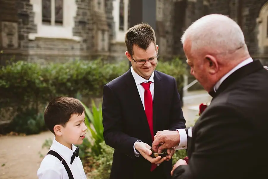 Boy in a bow tie standing beside two suited men outside a stone building.