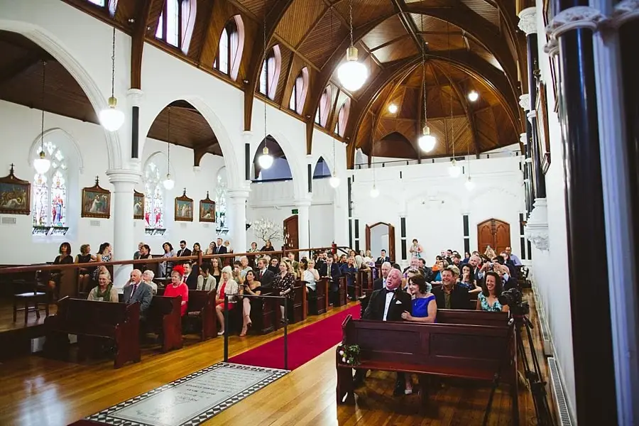 Wide view of seated wedding guests along a red-carpeted church aisle.