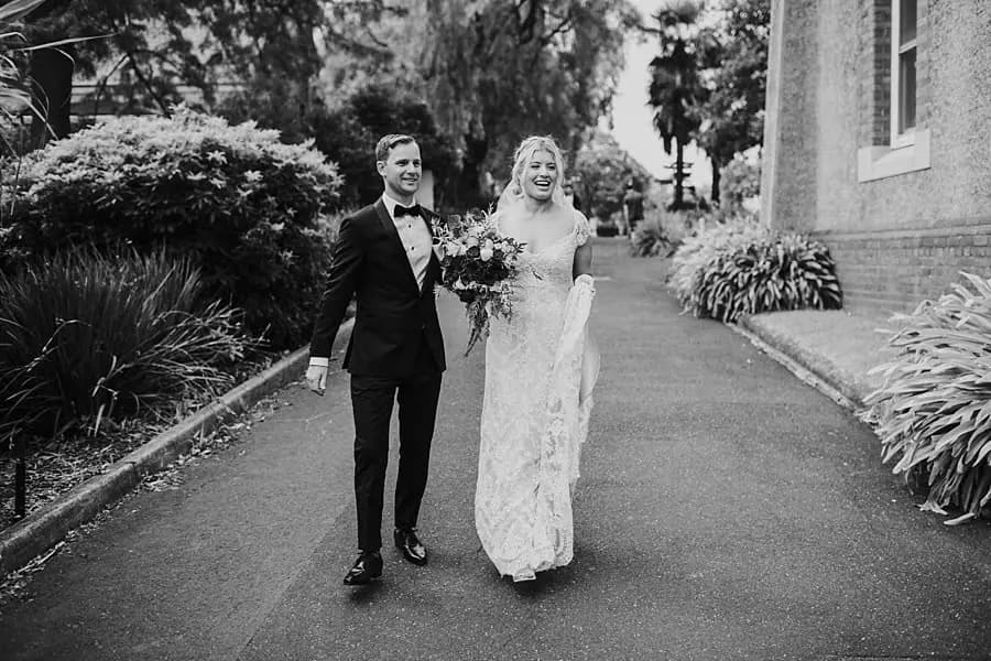 Black-and-white photograph of the bride and groom walking through leafy grounds.