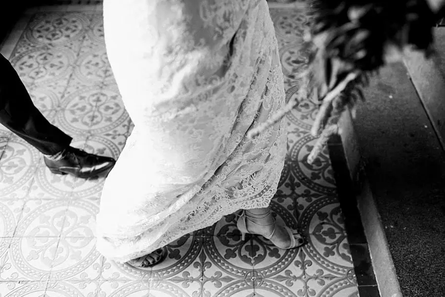 Black-and-white close-up of the bride's lace dress and shoes on patterned tiles.