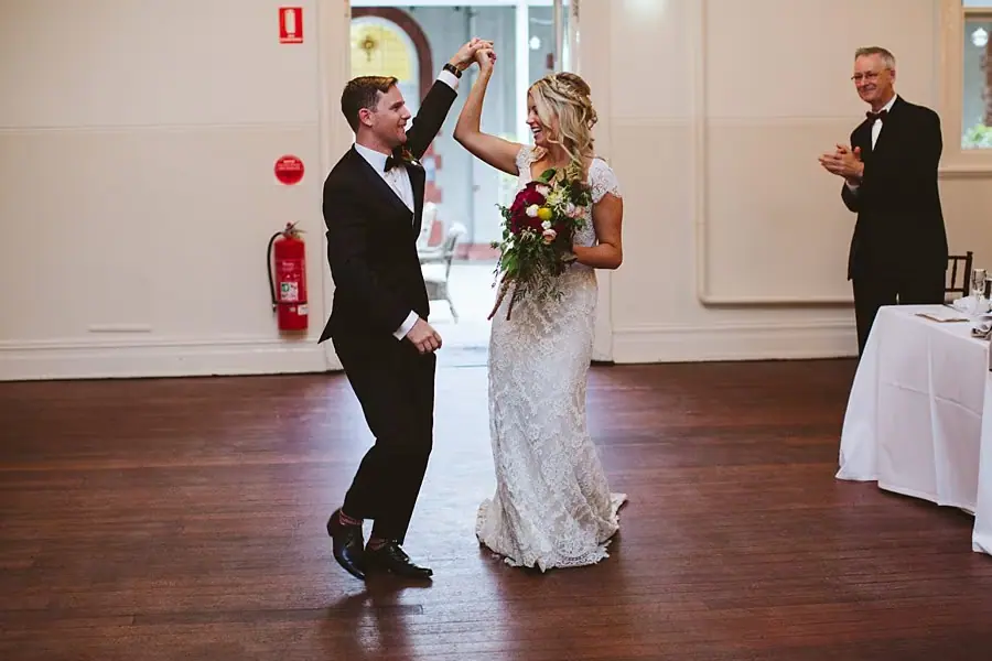Groom raising the bride's hand as they turn together on a wooden floor.
