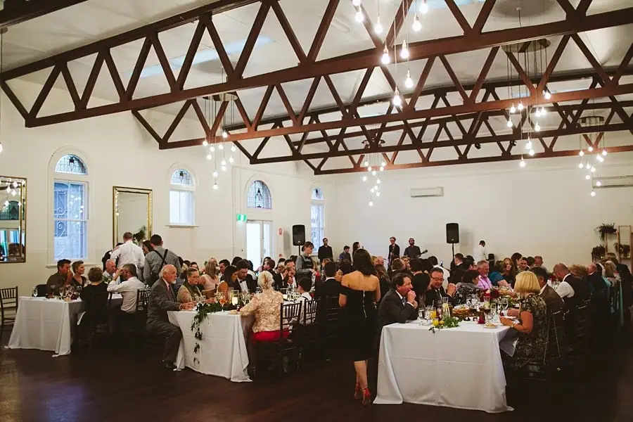 Wedding guests seated at reception tables beneath timber roof trusses.