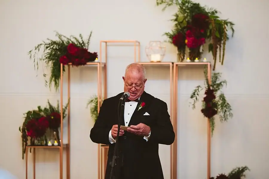 Suited man reading from a small card beside floral displays.
