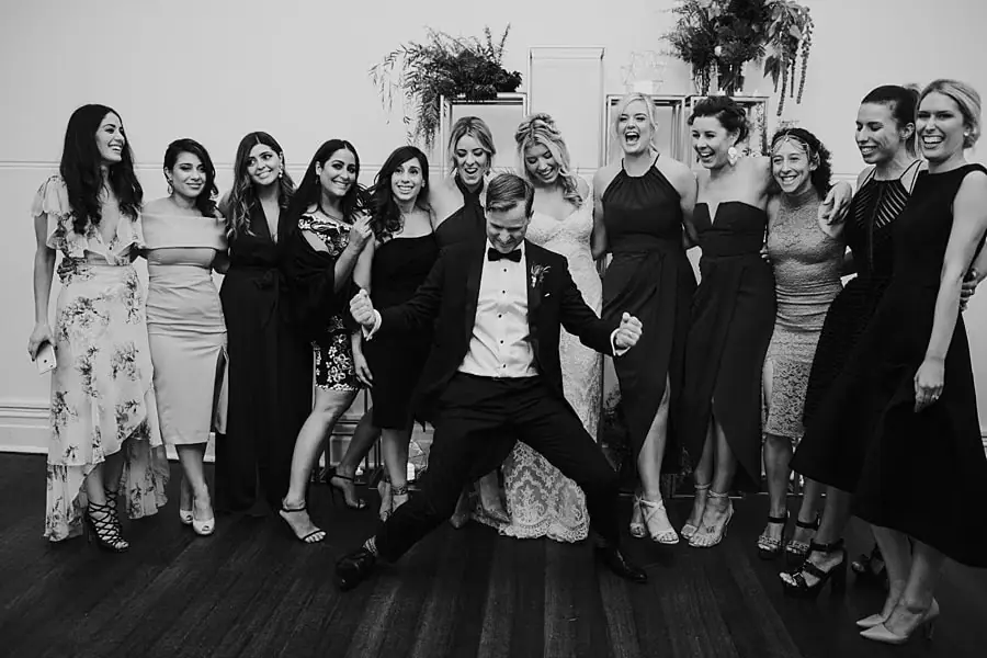 Black-and-white group photograph of women surrounding a seated man in a tuxedo.