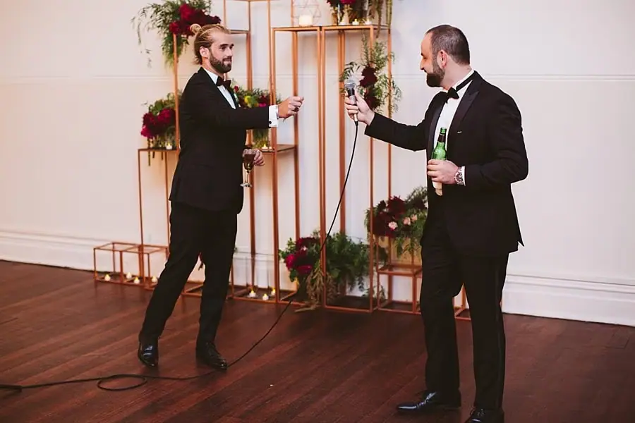 Two suited men speaking into microphones beside floral displays.