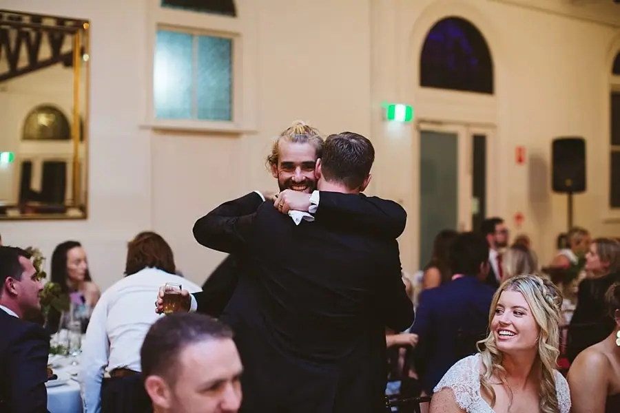 Two suited men embracing as the bride smiles beside them.