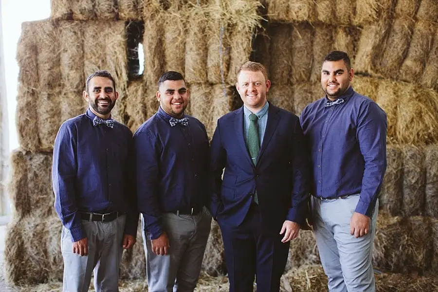 Groom and three groomsmen standing beside stacked hay bales