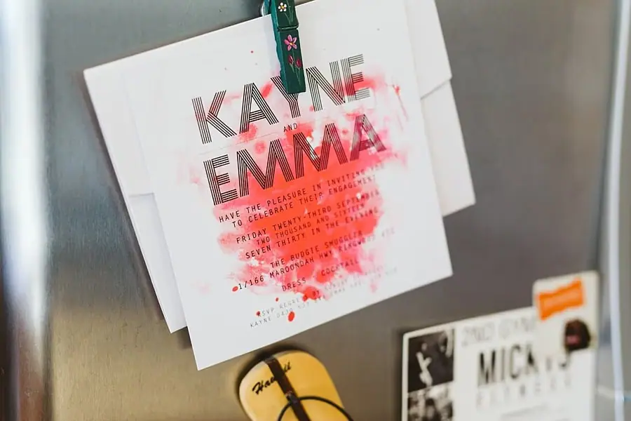 Colorful wedding invitation attached to a refrigerator with a clip.