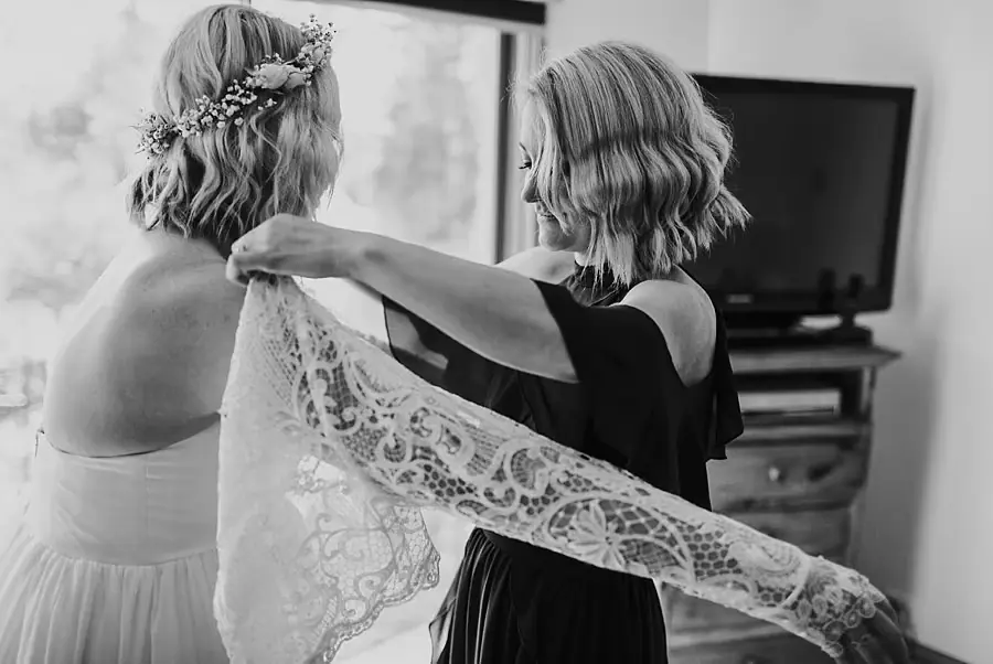 Black-and-white photograph of a woman helping the bride put on a lace top.