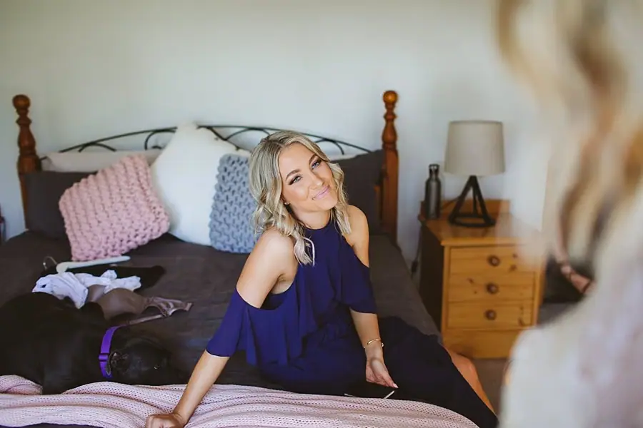 Woman in a purple dress smiling while seated on a bed.