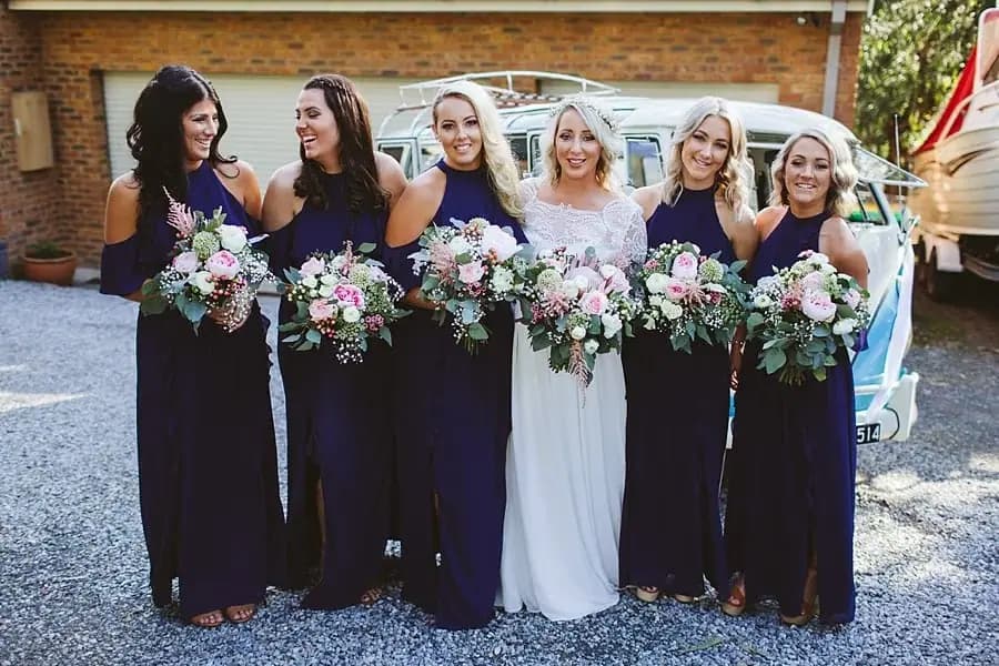 Bride and five women in purple dresses holding flower bouquets outside.