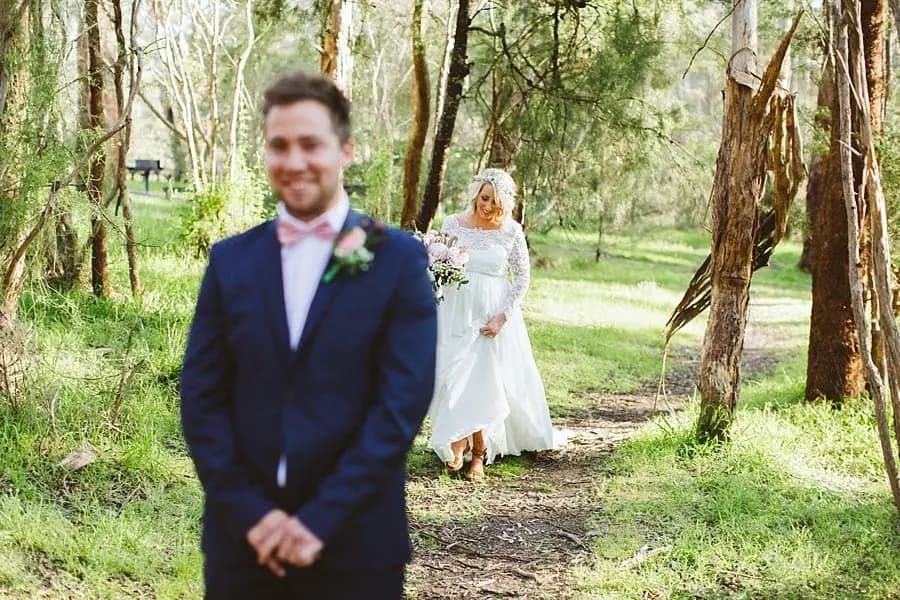 Groom standing in the foreground as the bride approaches through trees.
