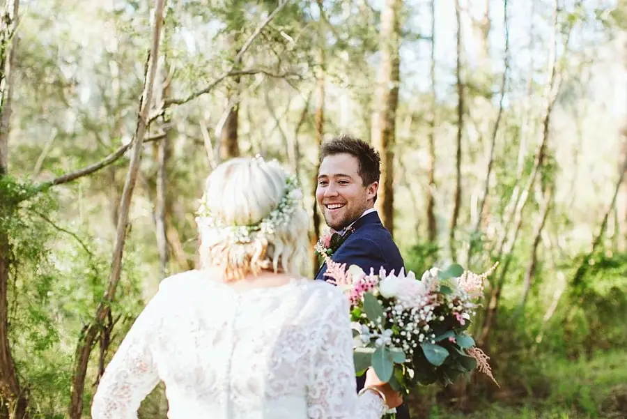 Groom turning and smiling toward the bride in woodland.