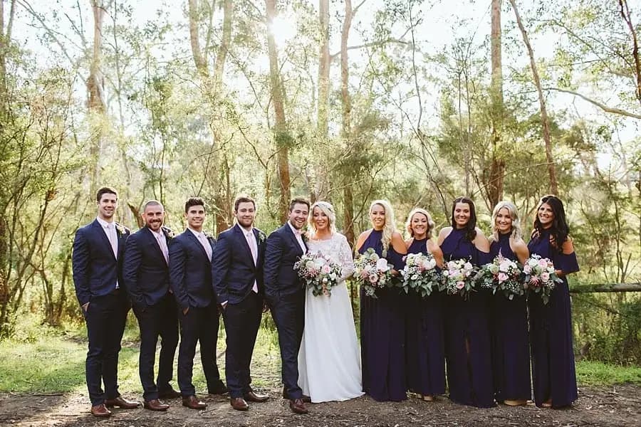 Wedding party posing in a line beneath tall trees.
