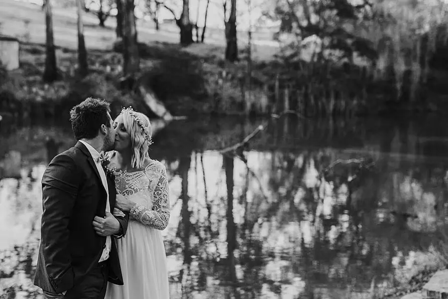 Black-and-white photograph of the couple kissing beside a reflective woodland pond.