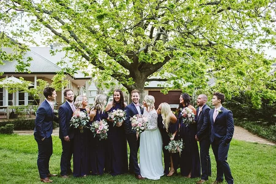 Wedding party gathering on a lawn beneath a leafy tree.