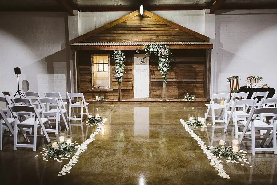 White ceremony chairs facing a flower-decorated timber structure indoors.