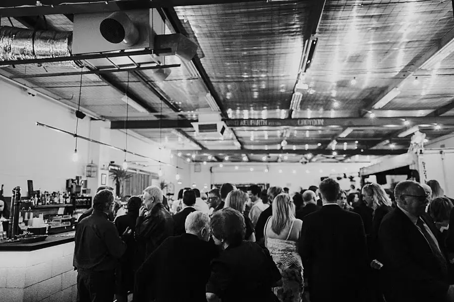 Black-and-white photograph of guests mingling beneath an industrial ceiling.