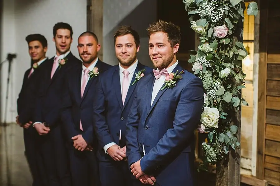 Groom and four suited companions standing beside a floral ceremony backdrop.