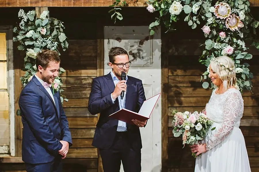 Bride and groom standing beside an officiant holding a microphone and folder.
