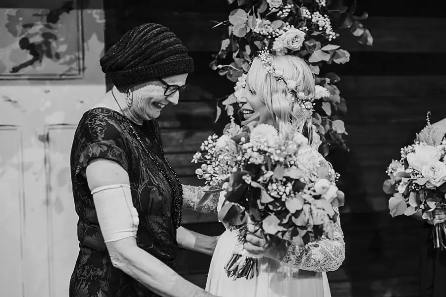 Black-and-white photograph of the bride embracing a woman beside ceremony flowers.