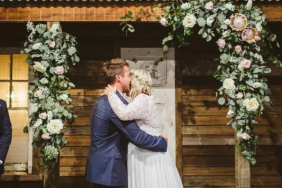 Bride and groom kissing beneath a flower-decorated timber backdrop.
