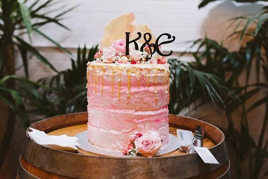 Pink two-tier wedding cake decorated with flowers and an initials topper.
