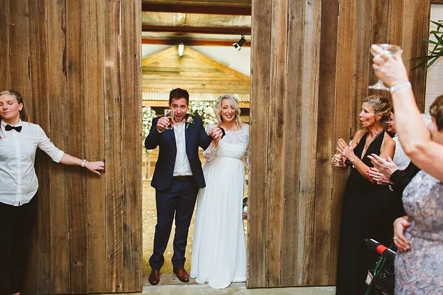 Bride and groom standing in a timber doorway as guests raise their glasses.