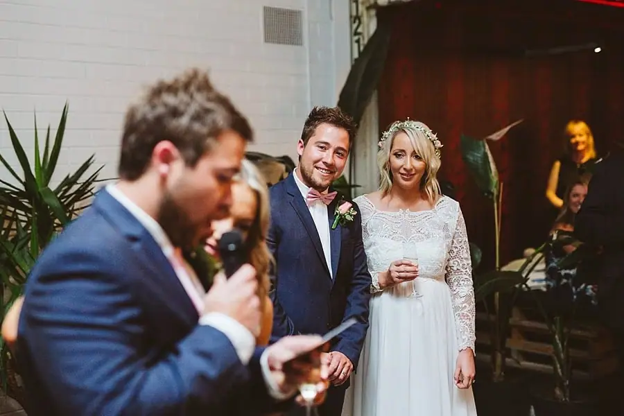 Suited man speaking into a microphone while the couple smiles behind him.