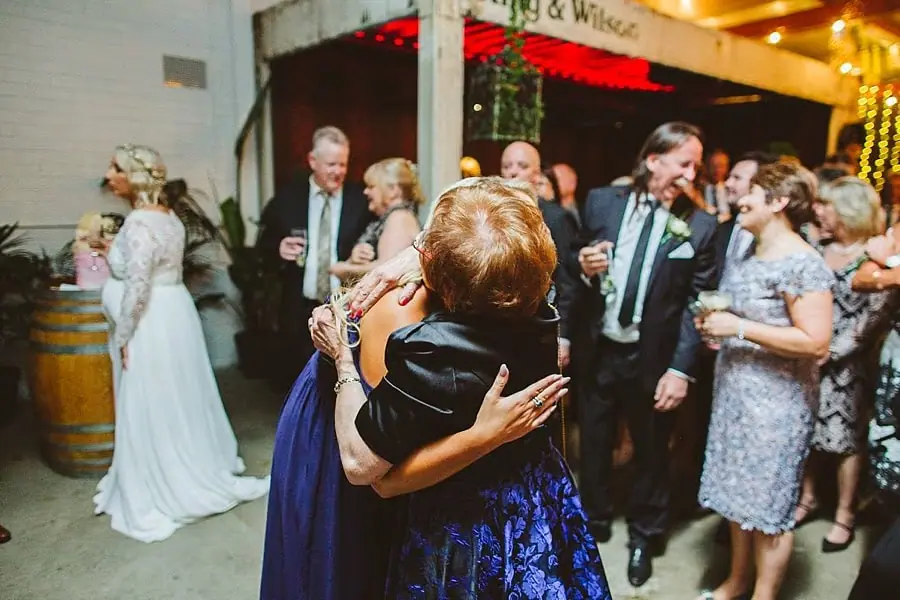 Two guests embracing in the foreground as reception guests chat behind them.