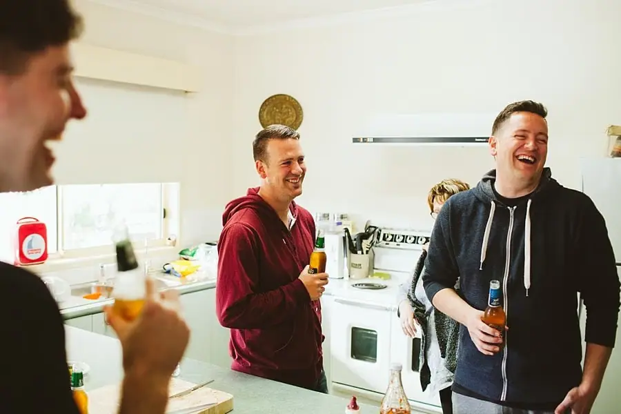 Men laughing together and holding drinks in a kitchen before the wedding.