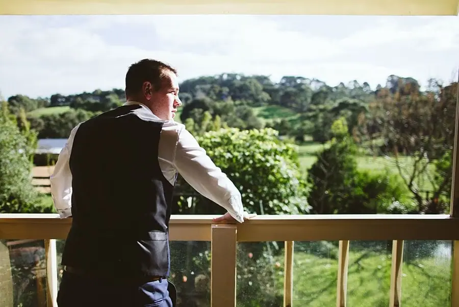 Man in a dark waistcoat leaning on a balcony railing overlooking green hills.
