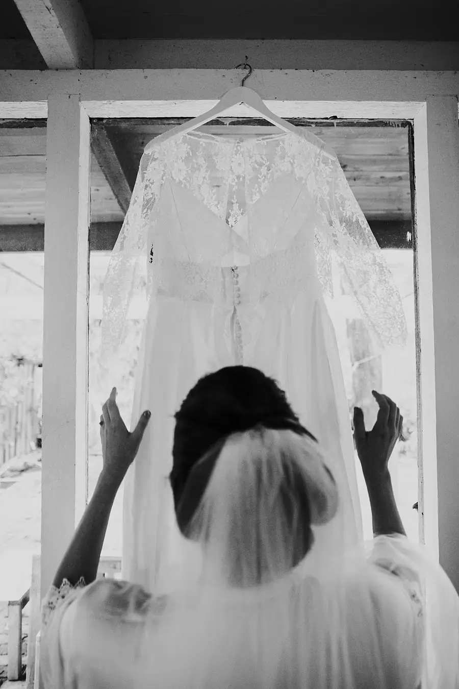 Black-and-white photograph of the veiled bride reaching toward a hanging wedding dress.