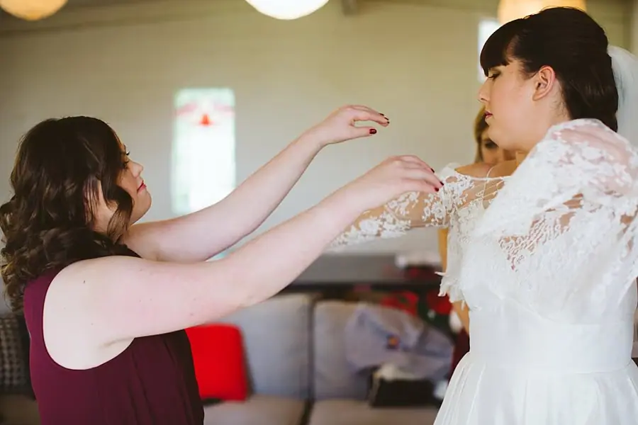 Woman in burgundy helping the bride adjust her lace wedding dress.