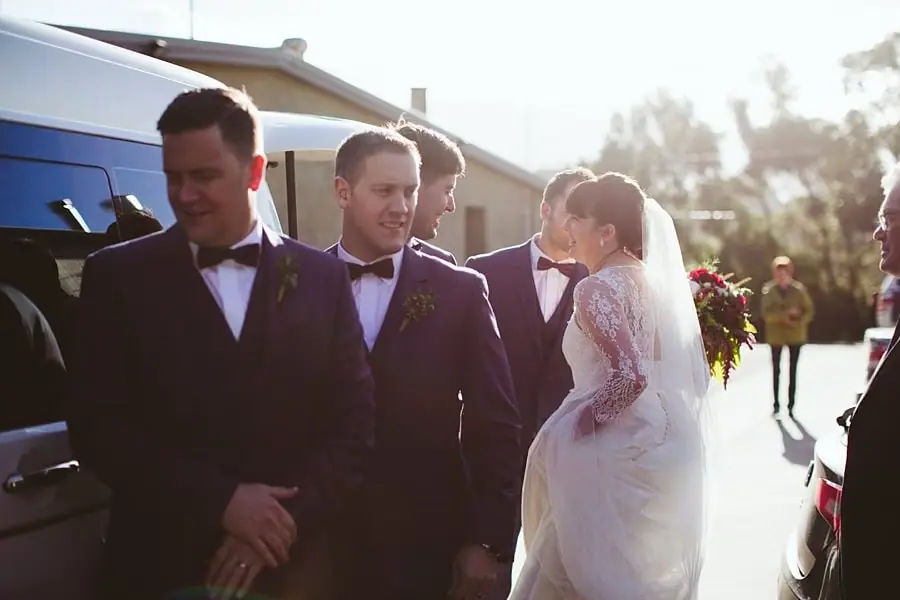 Bride and suited men gathering beside parked vehicles in the sunshine.