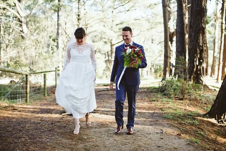 Bride lifting her dress as she walks barefoot beside the groom on a wooded path.