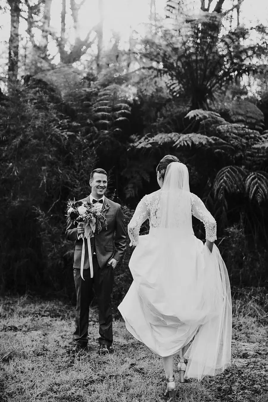 Black-and-white photograph of the bride lifting her dress while walking toward the groom in woodland.
