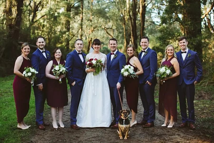 Wedding party in navy suits and burgundy dresses posing with the couple and a dog in woodland.