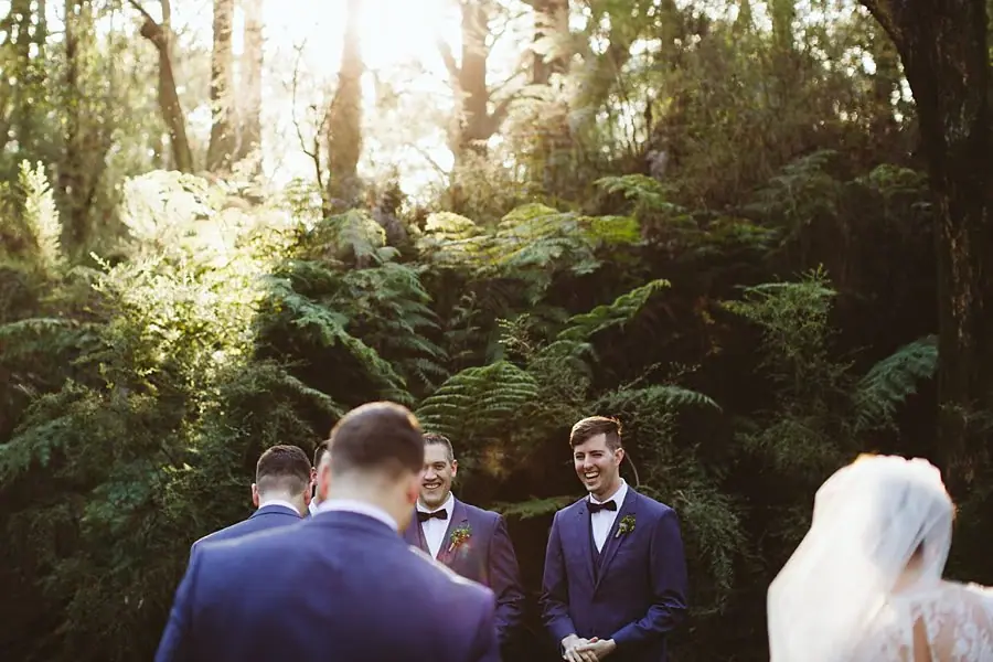 Suited men laughing together beneath sunlit trees as the bride stands nearby.