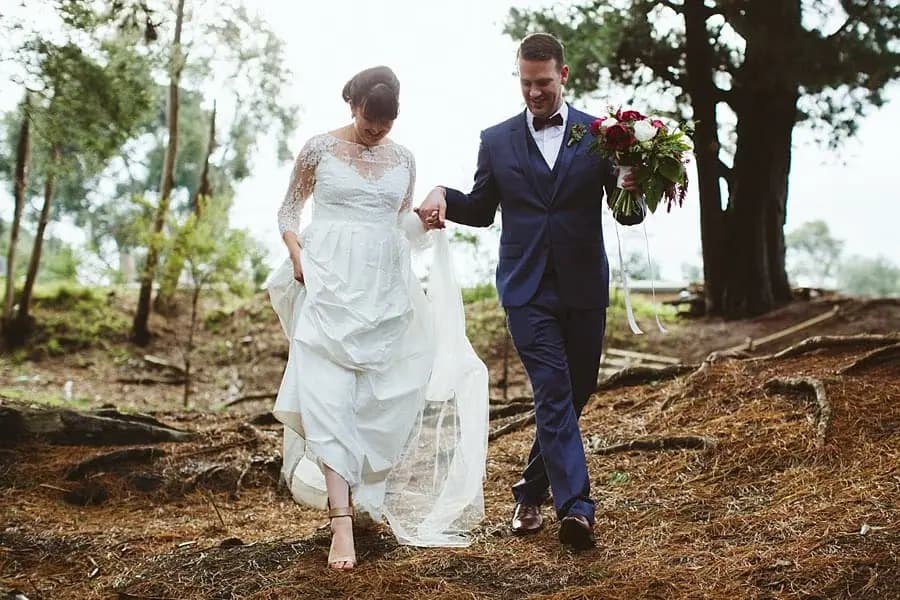 Groom holding the bride's hand as she walks barefoot down a wooded slope.