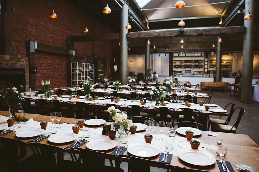 Reception tables set with plates, glasses, flowers and small potted succulents in a brick-walled room.