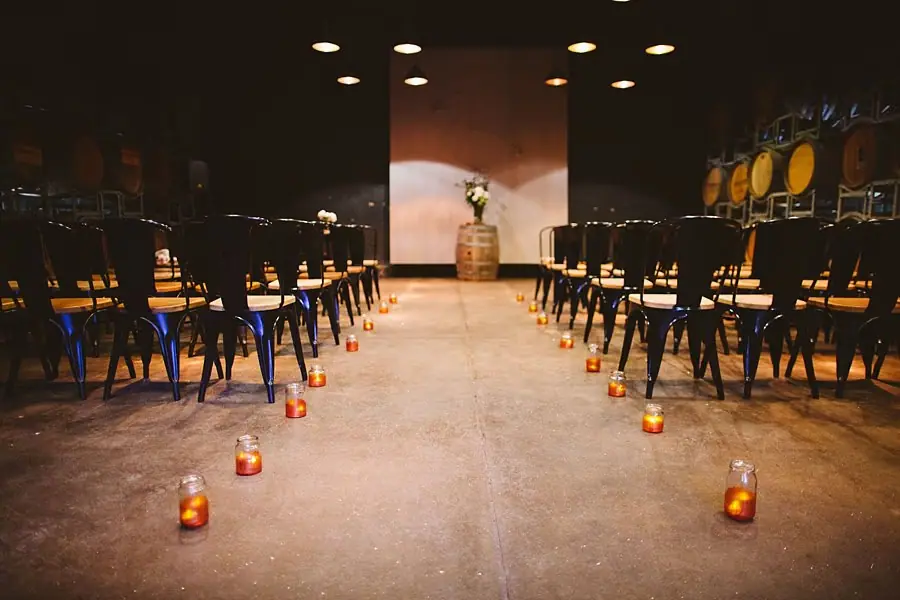 Candle-lined indoor ceremony aisle between rows of black chairs.